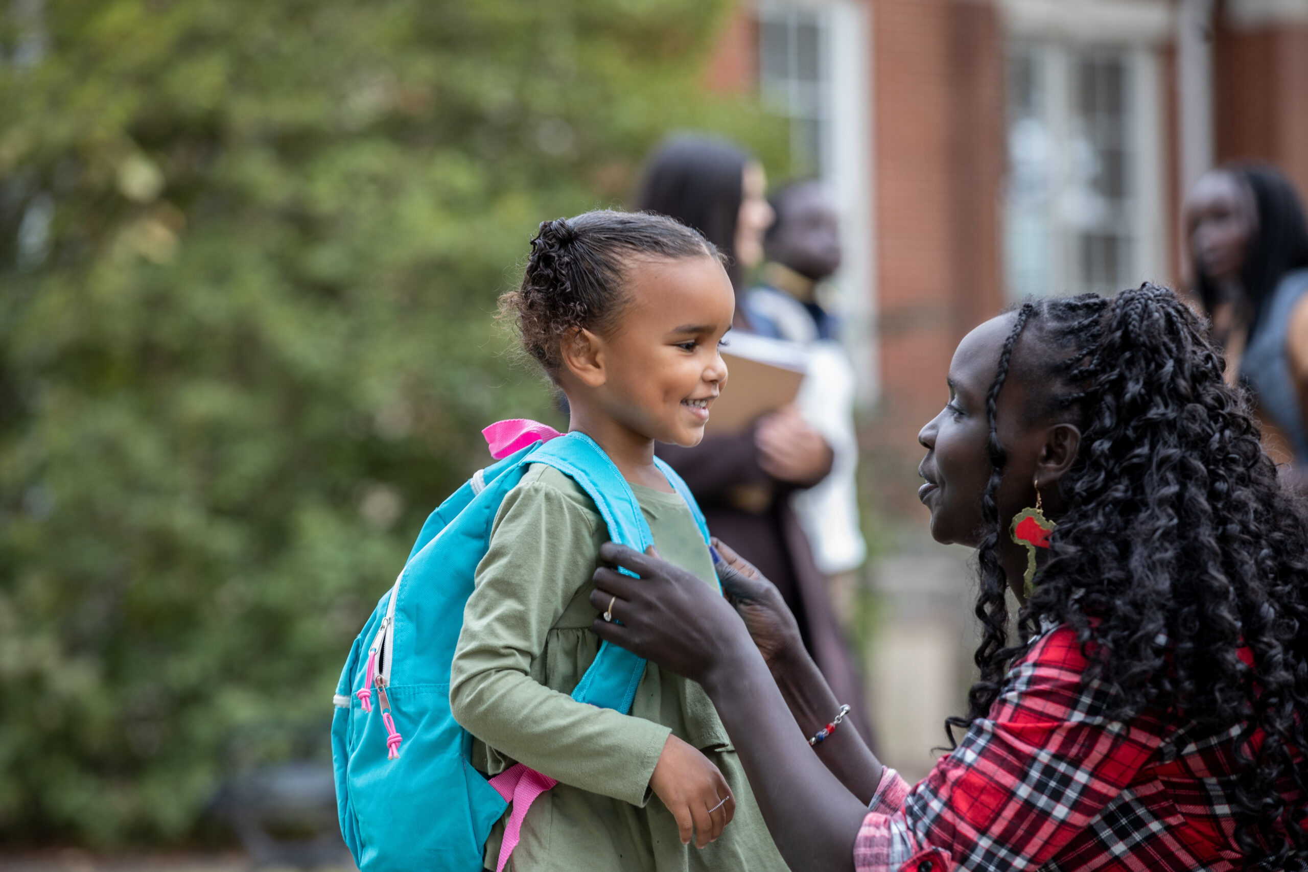 Mother kneels down to adjust backpack straps on small child's backpack in front of a school